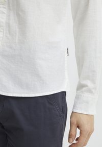 Biała lniana koszula z długim rękawem w stylu button-up, z zaokrąglonym dołem i małą metką marki z boku, w połączeniu z ciemnymi spodniami.
