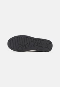 Semelle de chaussure en caoutchouc noir avec motifs antidérapants texturés et logo "HUGO" embossé au centre, affichée sur un fond clair uni.