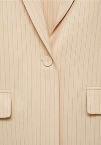 Blazer beige à fines rayures, fermeture par un seul bouton et deux poches avant à rabat visibles sur la partie inférieure de la veste.