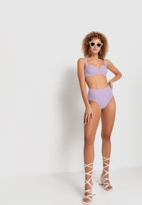 Lilac bikini top with underwire and adjustable straps, paired with high-waisted lilac bottoms. White strappy wedge sandals and square sunglasses.