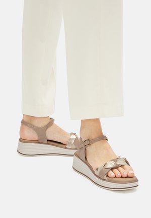 Beżowe sandały na koturnie z teksturowaną cholewką z tkaniny, metalicznym paskiem i plecionymi detalami. Posiadają pasek wokół kostki z klamrą w złotym odcieniu.