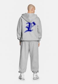 Szara bluza z dużym niebieskim logo na plecach, w zestawie z dopasowanymi szarymi spodniami dresowymi i białymi sportowymi butami. Prosty, luźny krój.