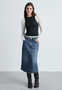 Zwart geribbeld mouwloos topje laagjes over een lange mouw witte shirt, gecombineerd met een high-waisted spijkerrok en kniehoge zwarte laarzen.