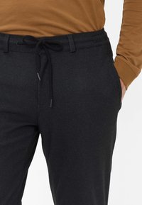 Zwarte textured broek met een broekband met trekkoord. Voorzien van zijzakken en een subtiel patroon. Gecombineerd met een bruine lange mouw shirt.