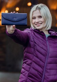 Navy leather wallet with a folded design and embossed logo, held in a hand. Background features warm lighting. Puffer jacket visible.