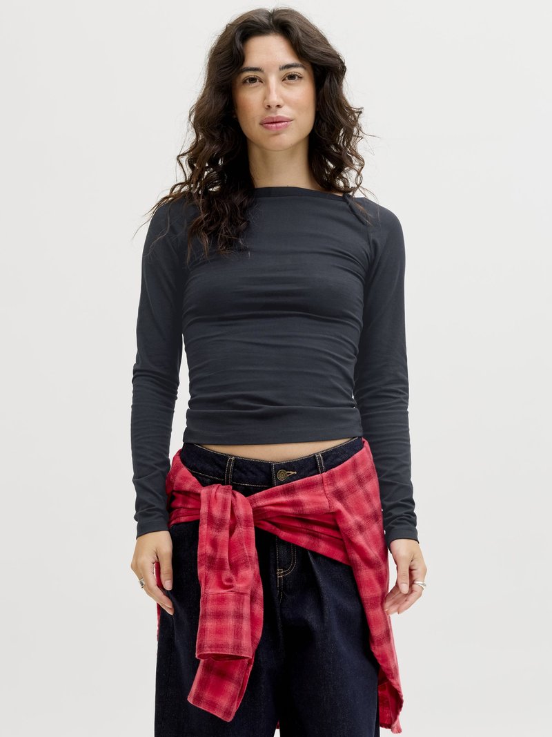 Femeie cu păr creț și întunecat, purtând un tricou negru cu mâneci lungi și blugi închiși la culoare, cu o cămașă roșie în carouri legată în jurul taliei.