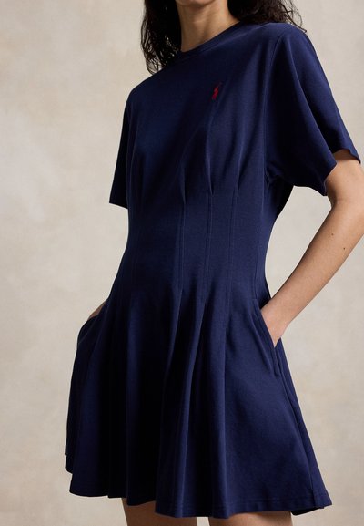 Frau trägt ein marineblaues Kleid mit kurzen Ärmeln, tailliertem Schnitt, ausgestelltem Rock, seitlichen Taschen und einem kleinen roten, aufgestickten Logo auf der Brust.