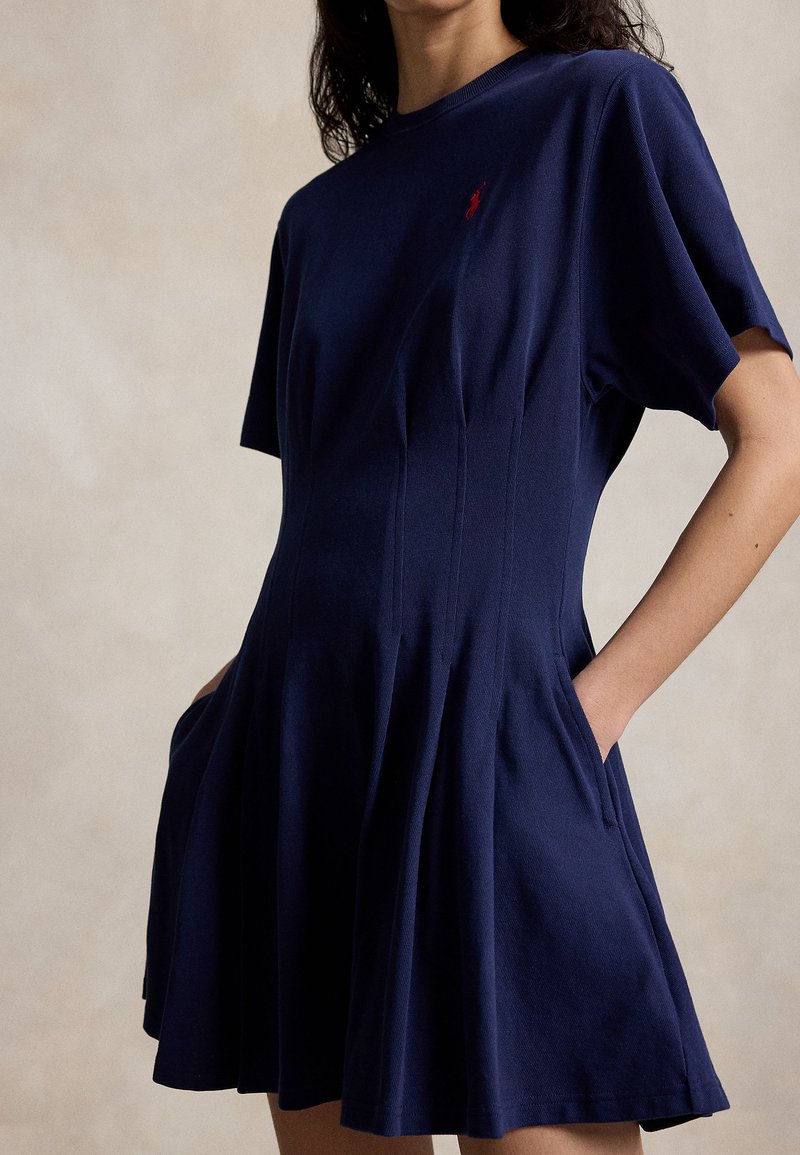 Frau trägt ein marineblaues Kleid mit kurzen Ärmeln, tailliertem Schnitt, ausgestelltem Rock, seitlichen Taschen und einem kleinen roten, aufgestickten Logo auf der Brust.