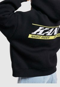Felpa nera con cappuccio e grafica sulla schiena. Presenta "KAN" in bianco, "NIGHT RIDER" in giallo e "EST 1989" in nero. Materiale morbido.