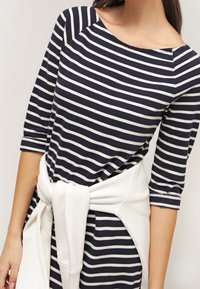 Femme portant une robe rayée bleu marine et blanche à manches trois-quarts, avec un pull blanc noué autour de la taille.