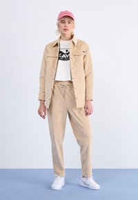 Beige kordfløyelsquiltet jakke over en hvit cropped skjorte med svart logo, kombinert med beige kordfløyelsbukser og hvite sneakers, toppet med en rosa caps.
