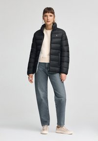 Czarna kurtka puchowa z wysokim kołnierzem, z pikowanymi sekcjami. Noszona na jasnoszary sweter i niebieskie dżinsy, w połączeniu z beżowymi trampkami.