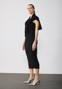 Nainen seisoo mustassa hihattomassa midi-mekossa, musta neule on heitetty hartioiden päälle ja hänellä on päällään hopeiset kärjelliset korkokengät.