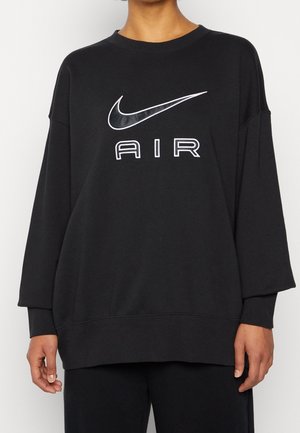 Osoba nosící volnou černou mikinu s bílým logem Nike ve tvaru háčku a nápisem "AIR" uprostřed na hrudi, doplněnou černými kalhotami.