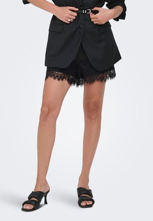 Kvinde iført sort blazer med bælte, sorte blonder shorts og sorte høje sandaler, står og justerer bæltet med hænderne.