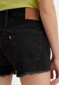 Pantalones cortos de mezclilla negra con dobladillo deshilachado, que cuentan con bolsillos traseros y una etiqueta de cuero marrón con "Levi Strauss & Co." en la cinturilla.