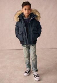 Chaqueta acolchada azul marino con capucha de piel sintética, bolsillos frontales, superpuesta a un suéter azul marino y una camisa de cuadros, combinada con jeans de mezclilla clara y zapatillas negras.