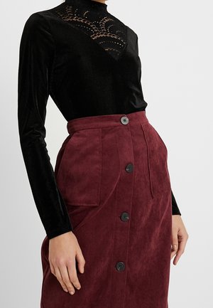 Top de terciopelo negro de manga larga con detalle de encaje en el cuello, combinado con una falda de pana burdeos con botones en la parte delantera y bolsillos grandes.