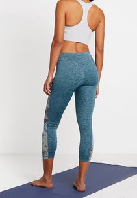 Kvinna som står barfota på en yogamatta, iförd en vit sportbh och blå mönstrade leggings, vänd bort från en enfärgad bakgrund.