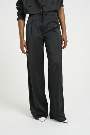 Pantalones negros de tiro alto con una tela suave y brillante. Presentan pierna ancha y pliegues, acompañados de zapatos blancos de punta.