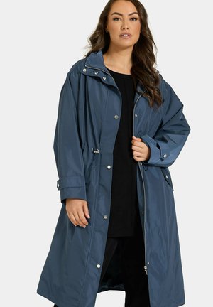 Donna che indossa un lungo impermeabile blu scuro sopra abiti neri, in piedi con una mano che tiene il cappotto vicino al petto.