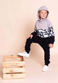 Szara bluza z kapturem z czarno-białym wzorem graficznym, zestawiona z czarnymi spodenkami dresowymi i białymi trampkami, chłopiec stojący na drewnianej skrzyni.