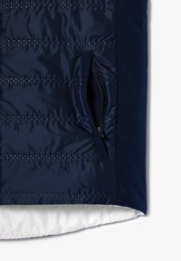 Chaleco acolchado azul marino con costuras horizontales, un bolsillo lateral con cremallera y un forro interior blanco en contraste. Textura suave y brillante.