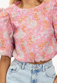 Blouse à motifs floraux dans des tons rose, orange et bleu avec un tissu texturé et des manches bouffantes, associée à un jean taille haute bleu clair.