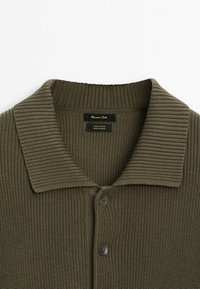 Cardigan en tricot côtelé vert olive avec un large col et deux boutons, étiquette indiquant 100 % coton, fabriqué en Chine.