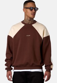 Brown sweatshirt with cream raglan sleeves, ribbed cuffs and hem. Features small white logo on chest. Styled with black pants.