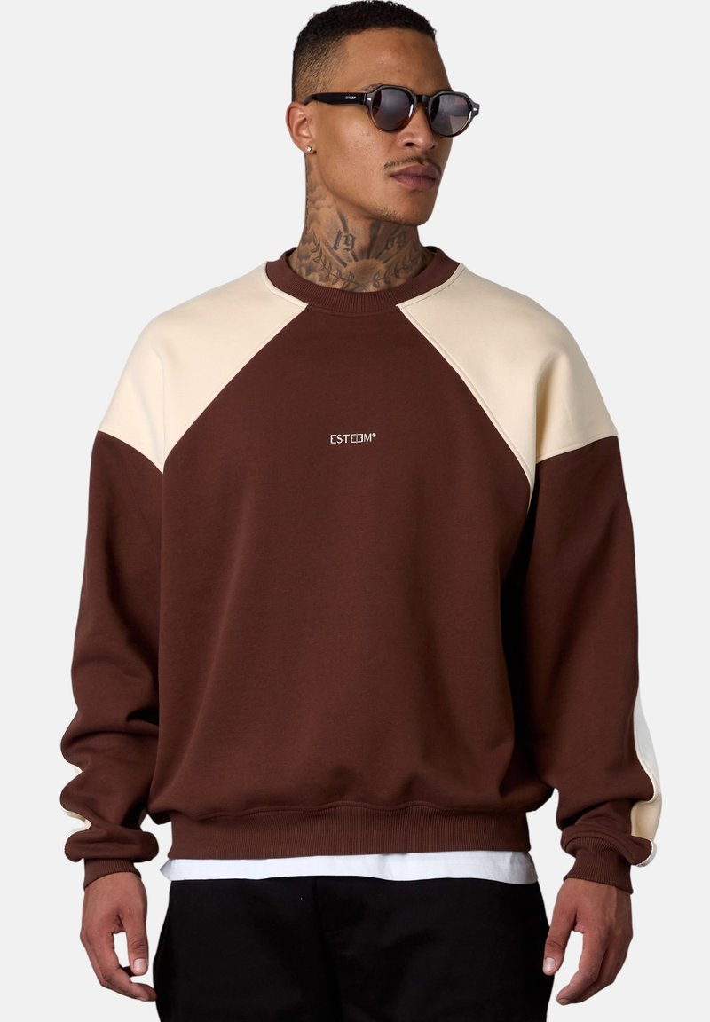Brown sweatshirt with cream raglan sleeves, ribbed cuffs and hem. Features small white logo on chest. Styled with black pants.