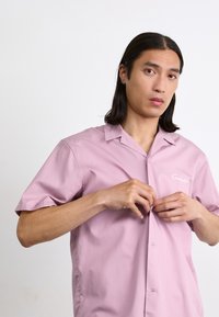Lichtroze T-shirt met korte mouwen, kampkraag en knoopsluiting. Heeft subtiele logo-geborduurd op de borstzak. Glad, lichtgewicht materiaal.