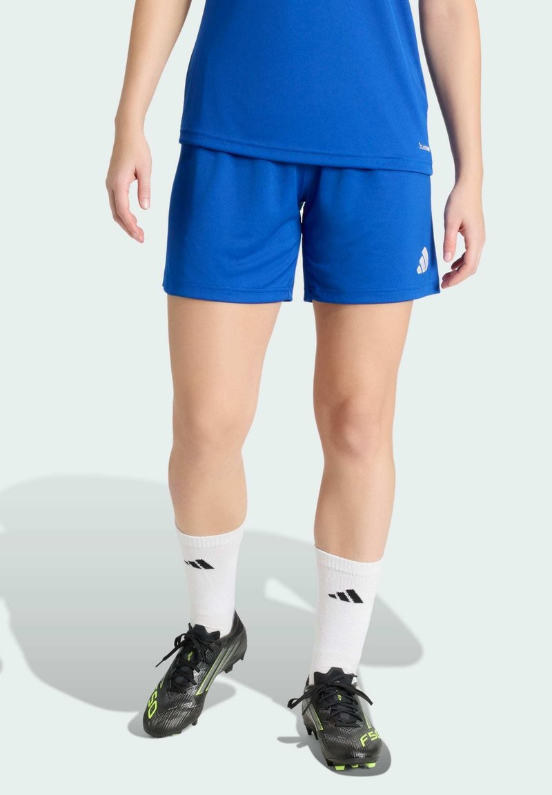 Personne portant un short de sport bleu, un t-shirt bleu assorti, des chaussettes blanches avec des logos noirs, et des chaussures à crampons noires, se tenant sur un fond clair.