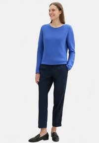 Mėlynas dryžuotas megztinis, tamsiai mėlynos spalvos pritaikytos kelnės ir juodi loaferiai. Megztinis turi apvalų iškirptę; kelnės tiesios formos su pilkais klostėmis.