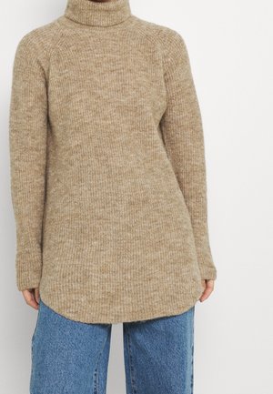 Beige stickad tröja med hög krage, långa raglanärmar och böjd nederkant. Texturen är mjuk och varm, med ett subtilt mönster.
