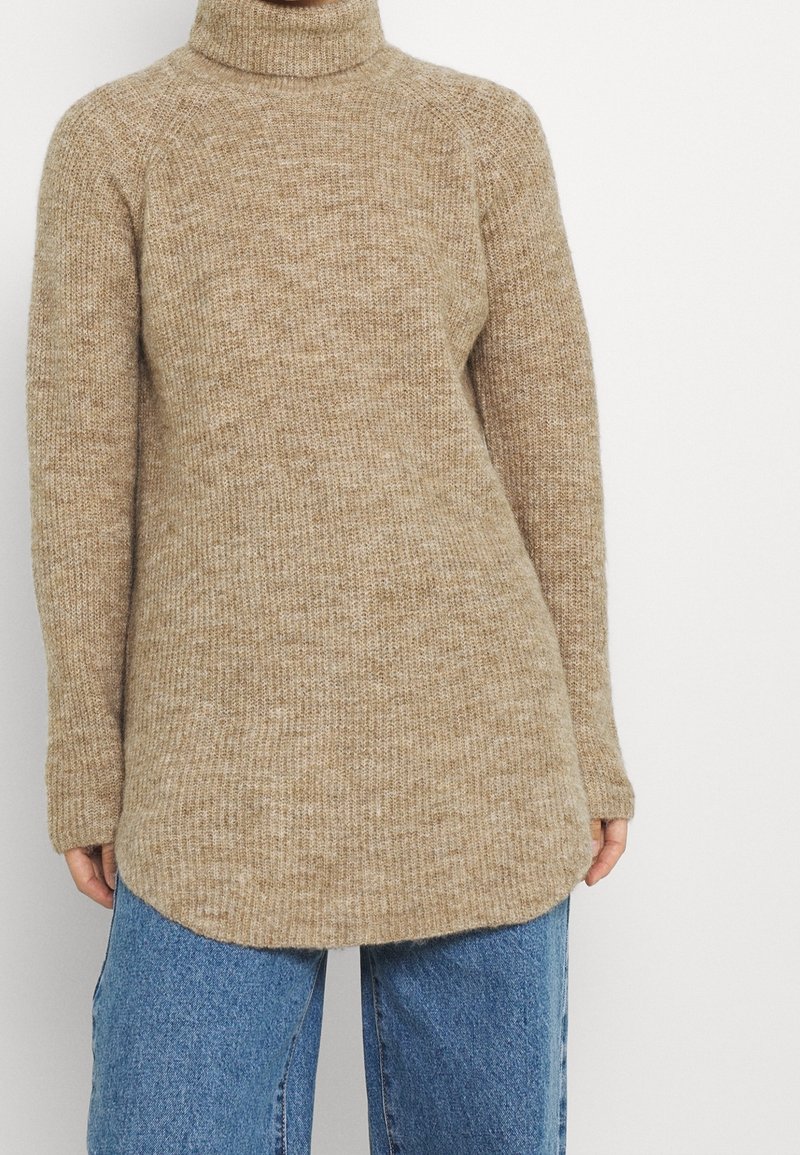 Suéter de punto beige con cuello alto, mangas raglán largas y dobladillo curvado. La textura es suave y cálida, con un patrón sutil.