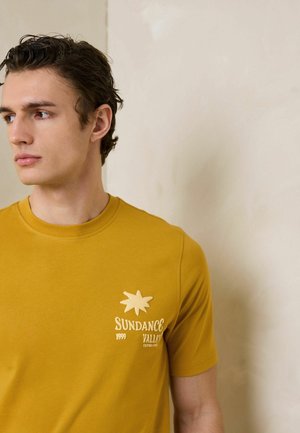 Tânăr purtând un tricou galben muștar cu inscripția "Sundance Valley 1999" și grafică cu soare, privind spre stânga pe un fundal neutru.