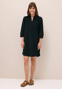 Tumma vihreä, polvipituinen mekko, jossa on kauluskotelokaulus ja kolmenneljänneksen hihat. Sileä rakenne, sivuleikkaukset ja hillitty nappidetaili kauluksessa.