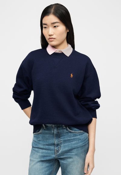 Γυναίκα με μακριά μαύρα μαλλιά, ντυμένη με μπλε σκούρο φούτερ πάνω από ένα ανοιχτό ροζ πουκάμισο με γιακά και μπλε τζιν, στέκεται μπροστά σε μια απλή φόντο.