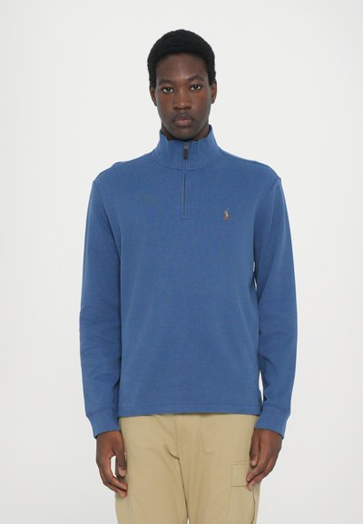 Blauwe fleece pullover met een hoge kraag en een halve rits, met een klein logo op de borst. Zachte textuur en een ontspannen pasvorm.