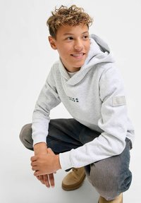 Sudadera gris claro con un logo frontal, bolsillo tipo canguro y puños acanalados. Combinada con jeans oscuros y botas beige, sobre un fondo neutro.