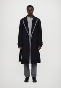 Trench coat in misto lana navy con dettagli grigi e design a doppiopetto, abbinato a un maglione a coste grigio chiaro e pantaloni grigi.