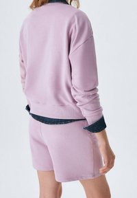 Person som bär en ljuslila sweatshirt och matchande shorts, med synliga rullade mörk denimskjortärmar under.