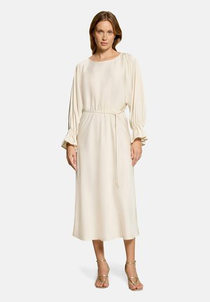Kvinde står med fronten frem iført en langærmet, off-white midi-kjole med bælte i taljen og guldfarvede strop-sandaler på ensfarvet baggrund.