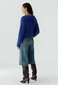 Frau von links mit einem flauschigen lila Pullover, verkürzten weit geschnittenen Jeans und knielangen, dunkelbraunen Stiefeln vor einem einfarbigen Hintergrund.