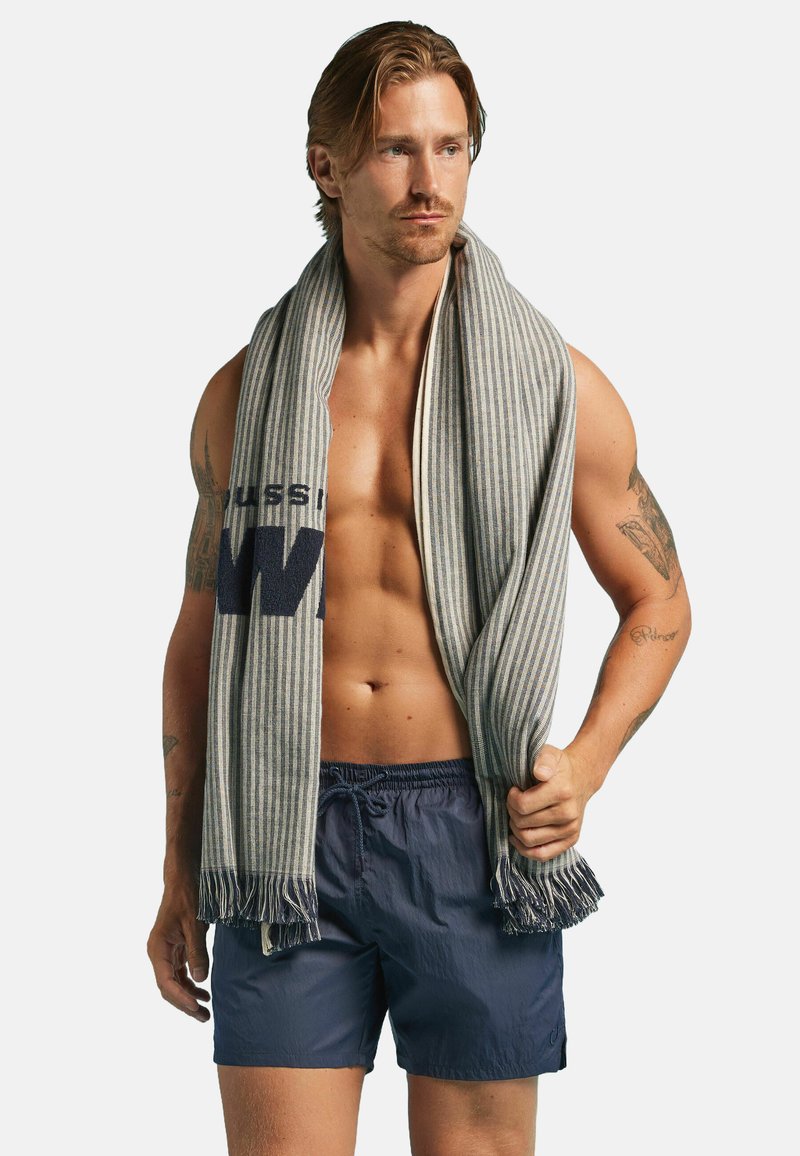 Serviette rayée en gris et bleu marin, avec des extrémités à franges. Modèle portant un short de bain bleu marin, tenant la serviette autour des épaules, avec un torse nu.
