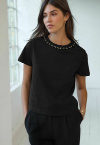 T-shirt noir à manches courtes avec un col rond agrémenté d'un design en chaîne. Le tissu semble lisse et léger.