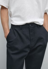 Pantaloni navy in cotone, con una texture liscia, tasche laterali, passanti per cintura e chiusura con bottone frontale. Abbinati a una camicia bianca.