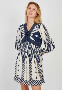 Korte mouw jurk in crème met marineblauwe geometrische patronen. Heeft een V-halslijn, koorddetail en een geplooide rok voor een ontspannen fit.