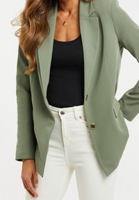 Blazer vert olive avec un design ajusté, simple boutonnage, deux boutons dorés, associé à un haut noir et un pantalon blanc.